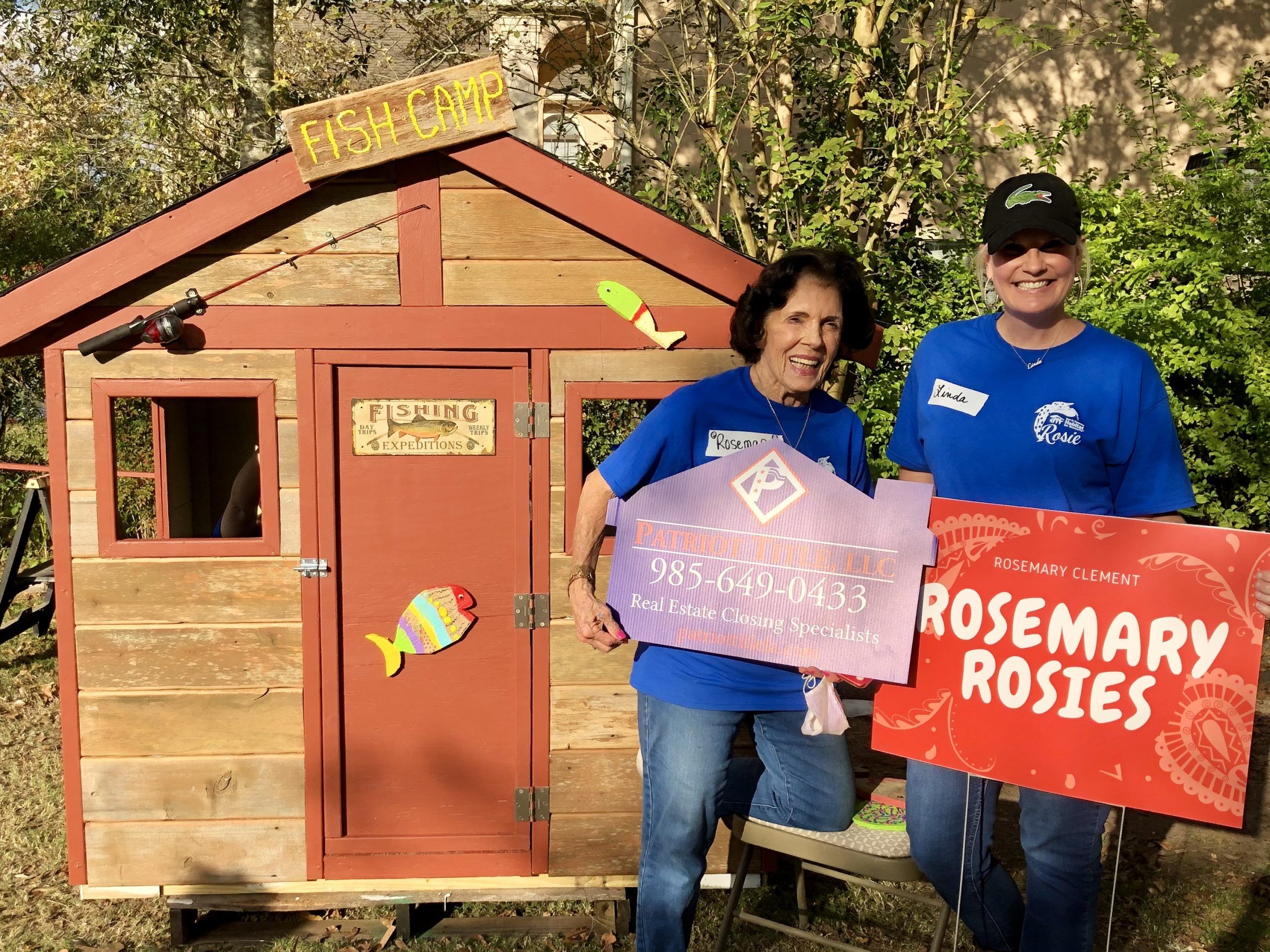 Two Patriot Title team members holding Patriot Title and Rosemary Rosies signs beside a children’s playhouse