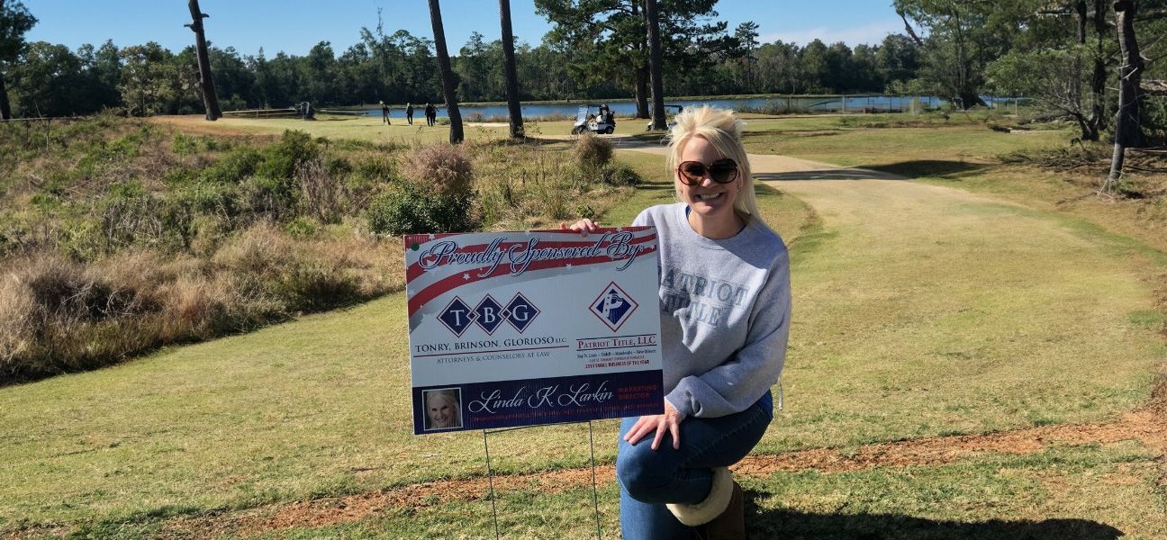 A Patriot Title team member kneeling beside a "Proudly Sponsored By" sign for Tonry, Brinson, Glorioso and Patriot Title on a golf course